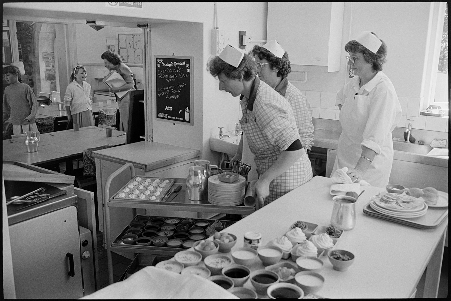 Primary school day. Teachers and pupils. Catering women making and serving lunch. Line up in yard.
[Three dinner ladies at Chulmleigh Primary School preparing to serve food in the canteen. One woman is pushing a trolley with various bowls and plates of food. Students and a teach can be seen in the dining room through the serving hatch.]