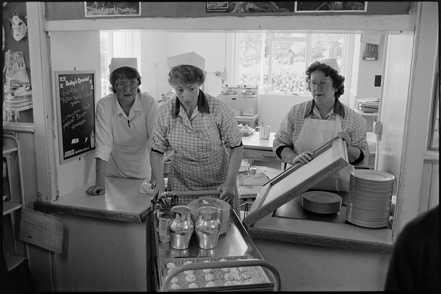 Primary school day. Teachers and pupils. Catering women making and serving lunch. Line up in yard.
[Three dinner ladies working in the canteen at Chulmleigh Primary School. One of them is holding up the serving hatch to let another woman push through a trolley with plates of food and jugs.]