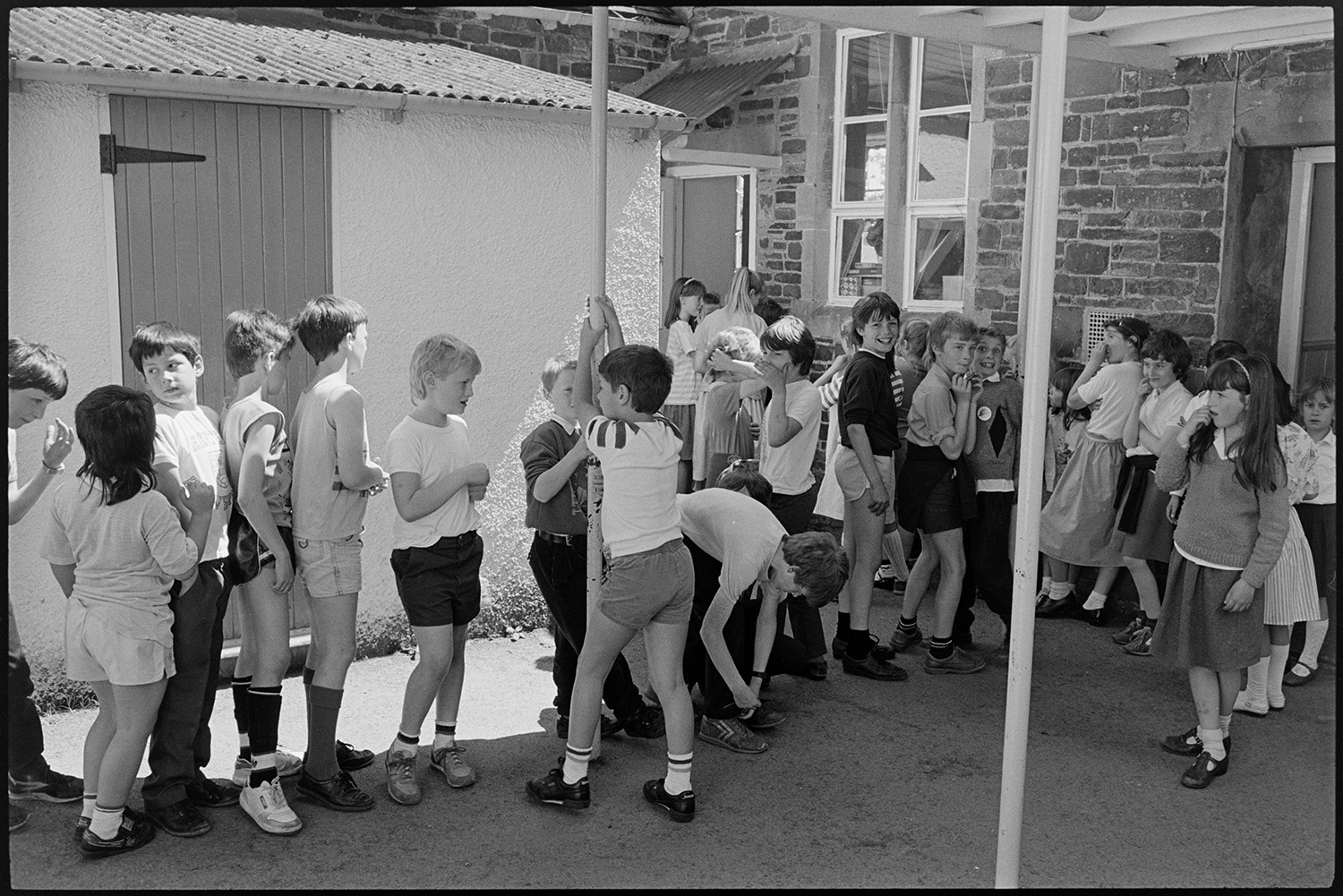 Primary school day. Teachers and pupils. Catering women making and serving lunch. Line up in yard.
[Children lining up in the playground and waiting near the doorway, possibly to the canteen, at Chulmleigh Primary School.]