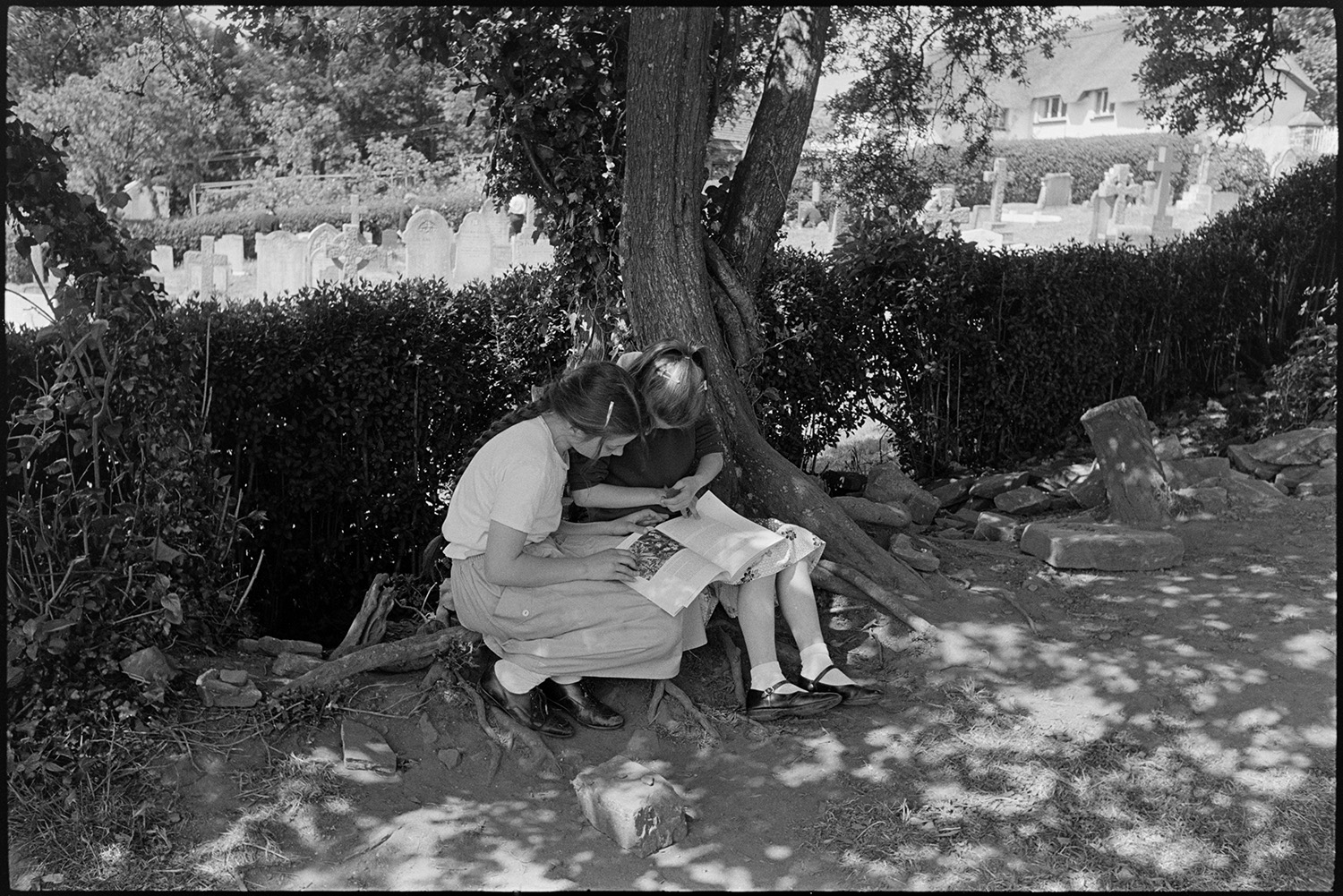A day in a primary school, cook making cakes, games in the playground, teaching,
[Two girls at Chulmleigh Primary School sitting under a tree and hedgerow next to the churchyard reading a book. A house with a thatched roof can be seen in the background.]