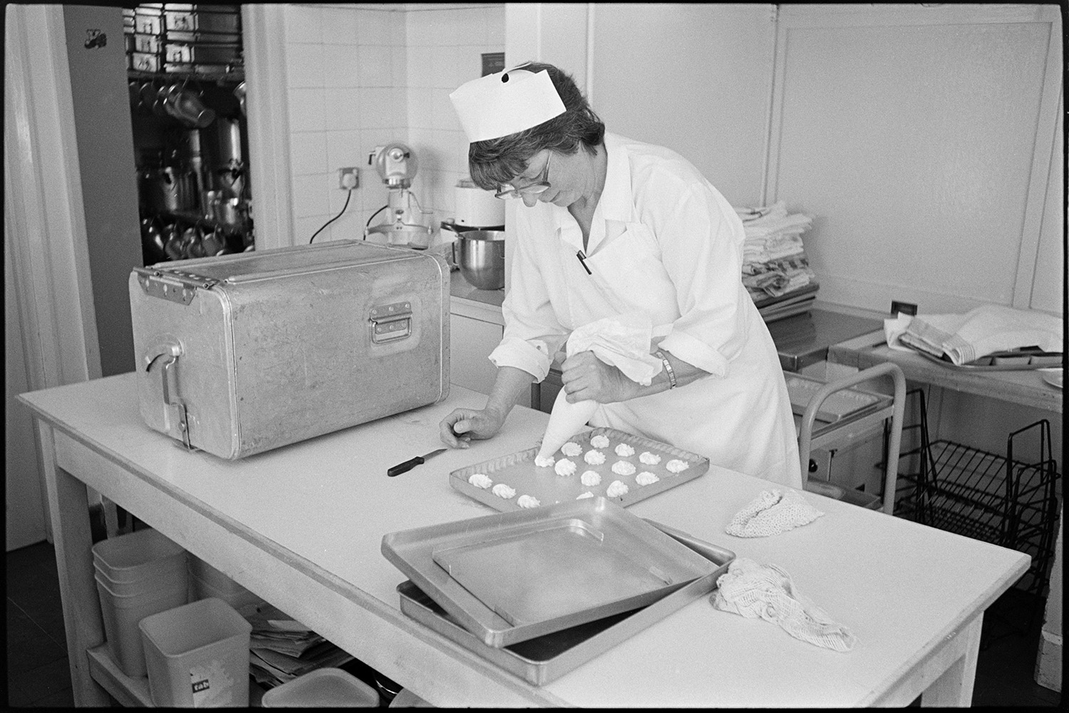 A day in a primary school, cook making cakes, games in the playground, teaching.
[A cook making cakes in the canteen at Chulmleigh Primary School. She is piping the mixture onto a baking tray. Trolleys and other cooking equipment is visible in the background.]