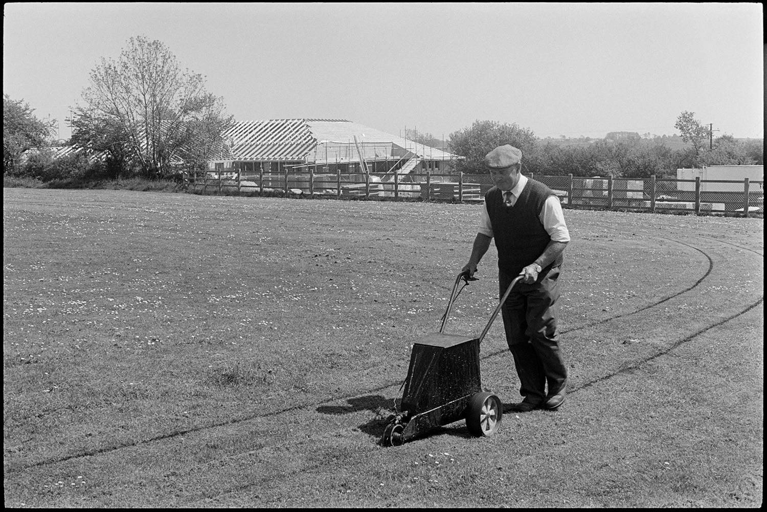 Man marking out school sports field.
[A groundsman marking a track around the edge of Chulmleigh Primary School sports field, pushing an line marking machine. In the background can be seen the construction of the new school building.]