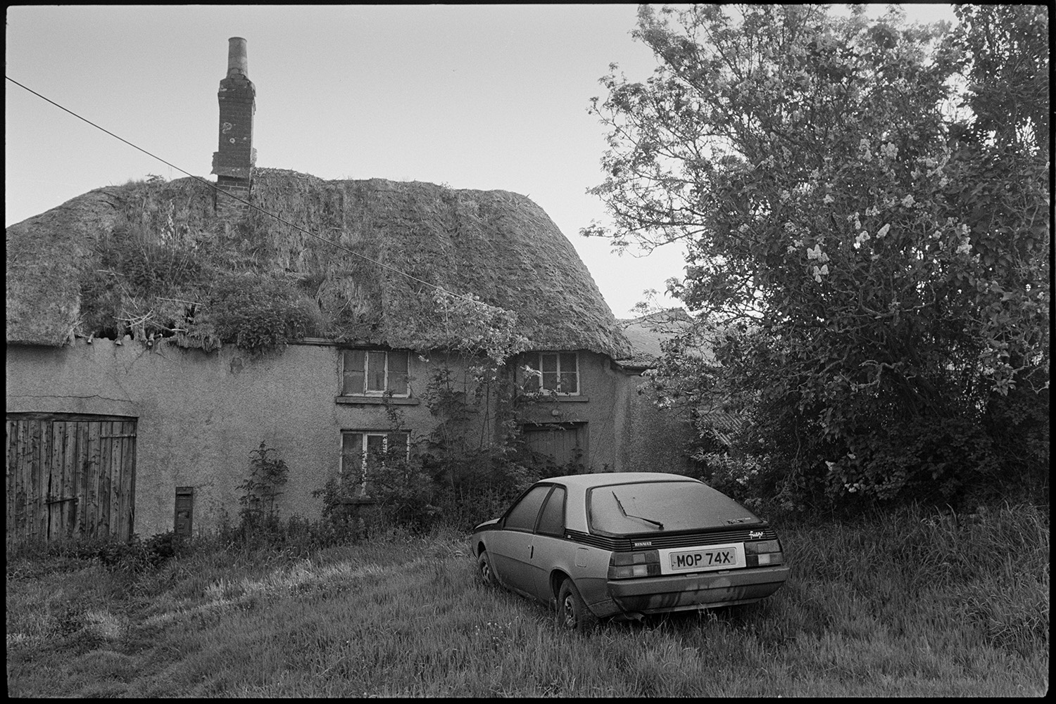 Derelict cob and thatch farm with letterbox in wall.
[An overgrown and derelict cob farmhouse with a thatched roof at Mary Week, Chulmleigh with a  Renault Fuego saloon car parked on the grass in front of the house. A wooden garage door and post box can be seen in the wall of the house.]