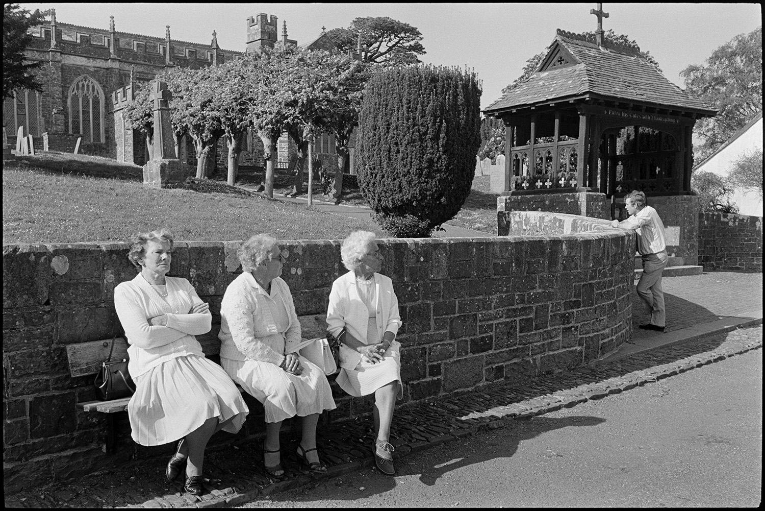 Three women on bench in front of church gate.
[Three women sitting on a bench and a man leaning on the wall in front of the lychgate entrance to Chittlehampton Church. An avenue of trees leads up to the church.]