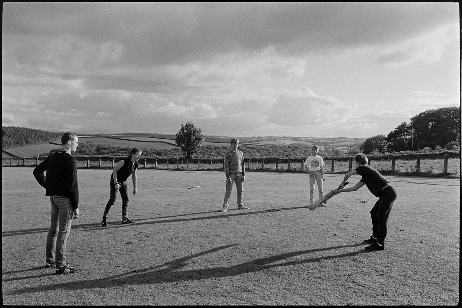 Boys cricket fielding practice on village cricket pitch with sun and clouds.
[Five boys at cricket practice with one of the boys hitting the ball with a bat, on the Chulmleigh village cricket field. A view of the surrounding countryside of fields and hedgerows is visible in the background along with clouds in the sky.]