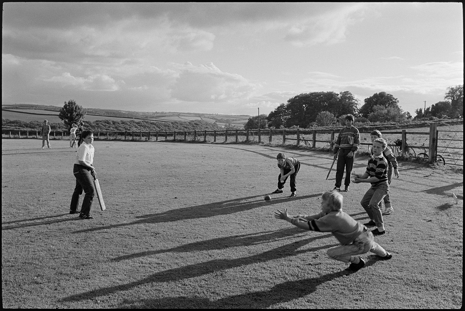 Boys cricket fielding practice on village cricket pitch with sun and clouds.
[A group of five boys at cricket practice, with one boy holding a bat and one boy diving to catch the ball. They are being coached by Mr Parker on the Chulmleigh village cricket field. A view of the surrounding countryside of fields and hedgerows is visible in the background along with clouds in the sky.]