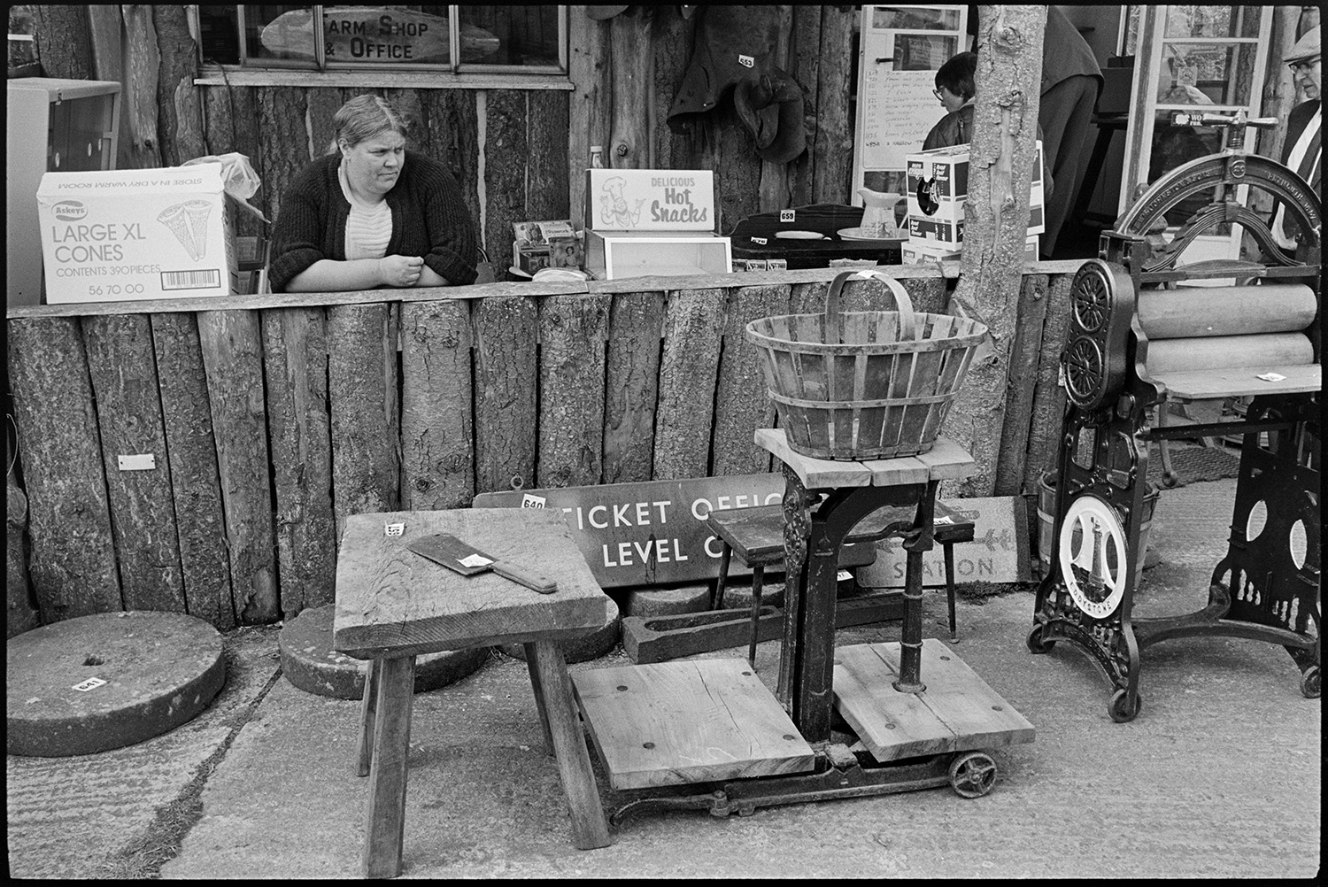 Sale of agricultural museum collection, signs, implements, machinery on display. Woodpile.
[A woman selling ice creams and hot snacks at a counter at the Ashley Collection, Hollocombe. In front of the counter are a number of items for sale, including grinding stones, a stool, trolley, basket, hand operated mangle or wringer and a railway Ticket Office sign.]