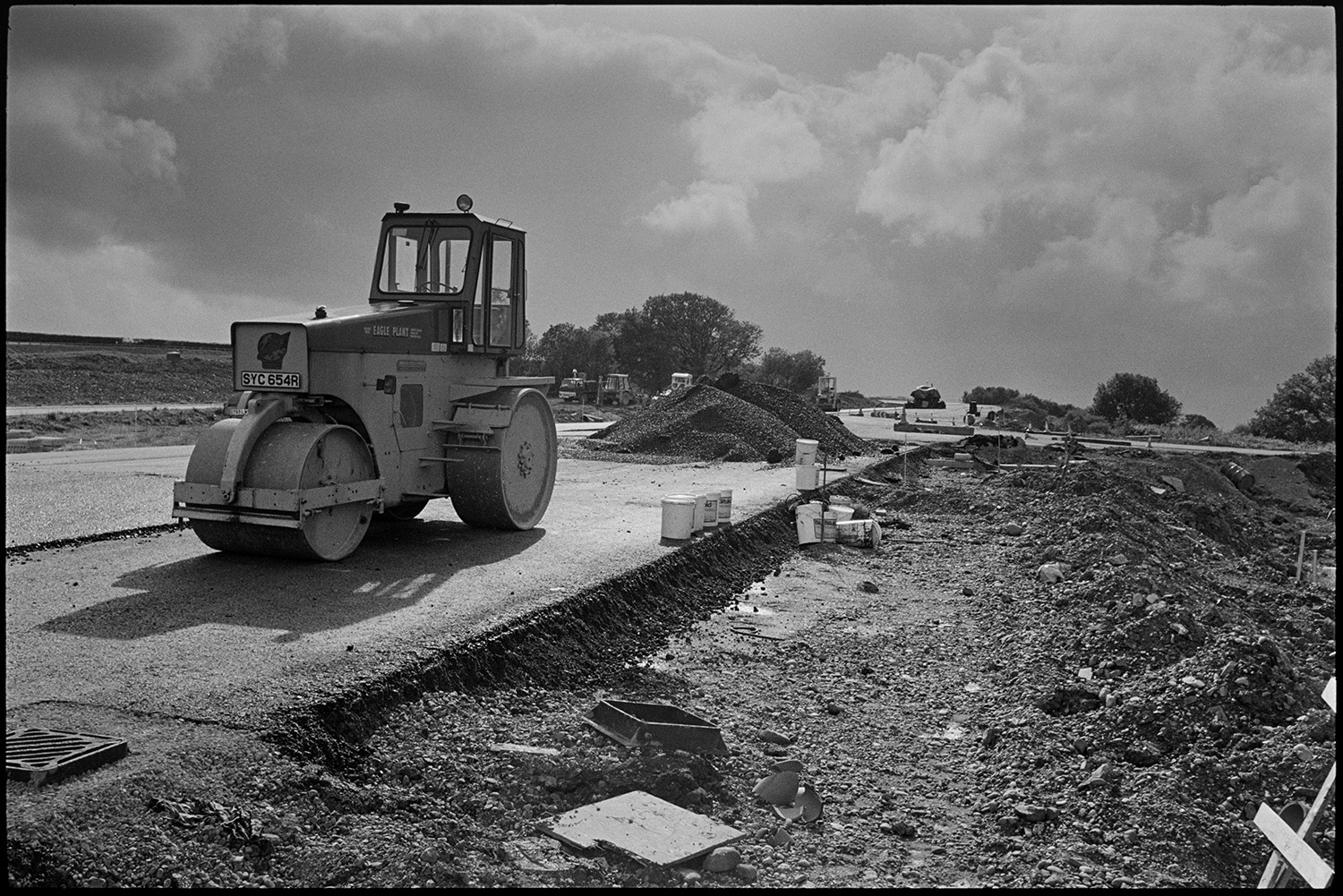 Machinery on new motorway link road shortly before it opened. Diesel roller and earth mover.
[A diesel roller working on the North Devon link road near Rackenford shortly before it opened. Other machinery, piles of gravel and buckets are visible on the road. A stormy sky is in the background.]