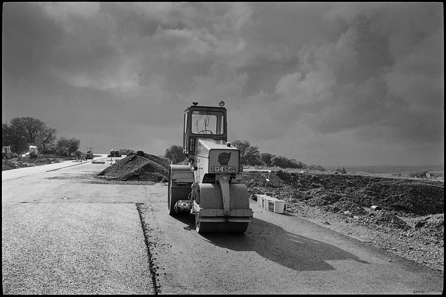 Machinery on new motorway link road shortly before it opened. Diesel roller and earth mover.
[A diesel roller working on the North Devon link road near Rackenford shortly before it opened. Other machinery, piles of gravel and buckets are visible on the road. A stormy sky is in the background.]