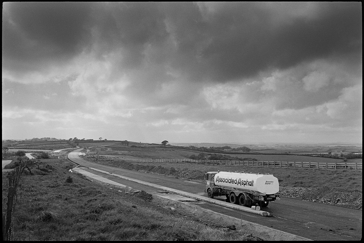 Standing stone beside new road it was moved before the road was laid down, passing tanker.
[A tanker lorry belonging to Associated Asphalt travelling along the new North Devon link road near Rackenford just before it opened. There is a wooden fence and rural landscape with fields and hedgerows in the background, with a stormy sky above.]