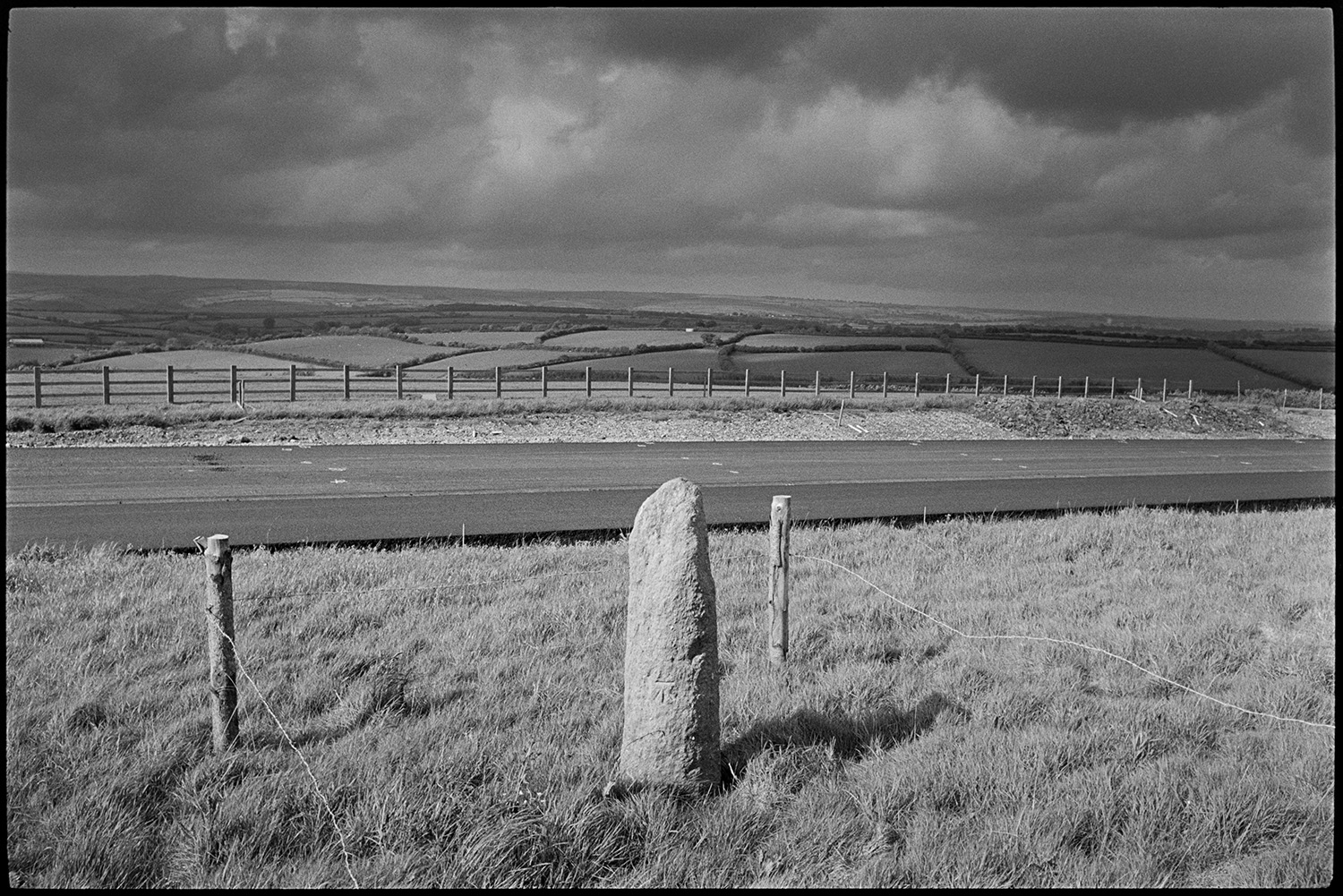 Standing stone beside new road, it was moved before the road was laid down, passing tanker.
[An old standing stone, fenced off by posts and barbed wire, alongside the new North Devon link road near Rackenford. There is a wooden fence and rural landscape of fields and hedgerows in the background, with a stormy sky above.]