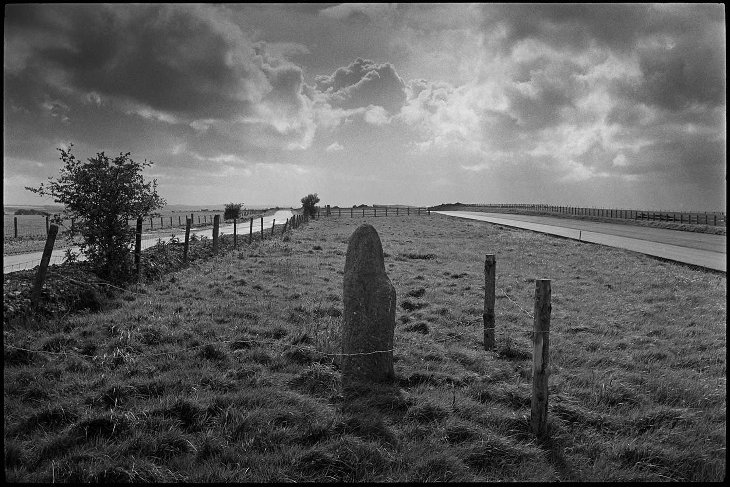 Standing stone beside new road, it was moved before the road was laid down, passing tanker.
[An old standing stone, fenced off by posts and barbed wire, alongside the new North Devon link road near Rackenford. There is another smaller road and  wooden fences in the background, with a stormy sky above.]