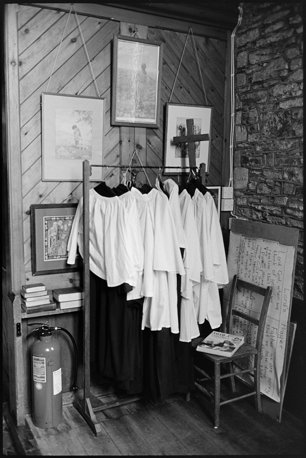 Clergy or choir robes hung on a rail in a church vestry. Also shown is a fire extinguisher, a chair, books, a pin board, a wooden cross, and pictures hanging on a wood panelled wall.