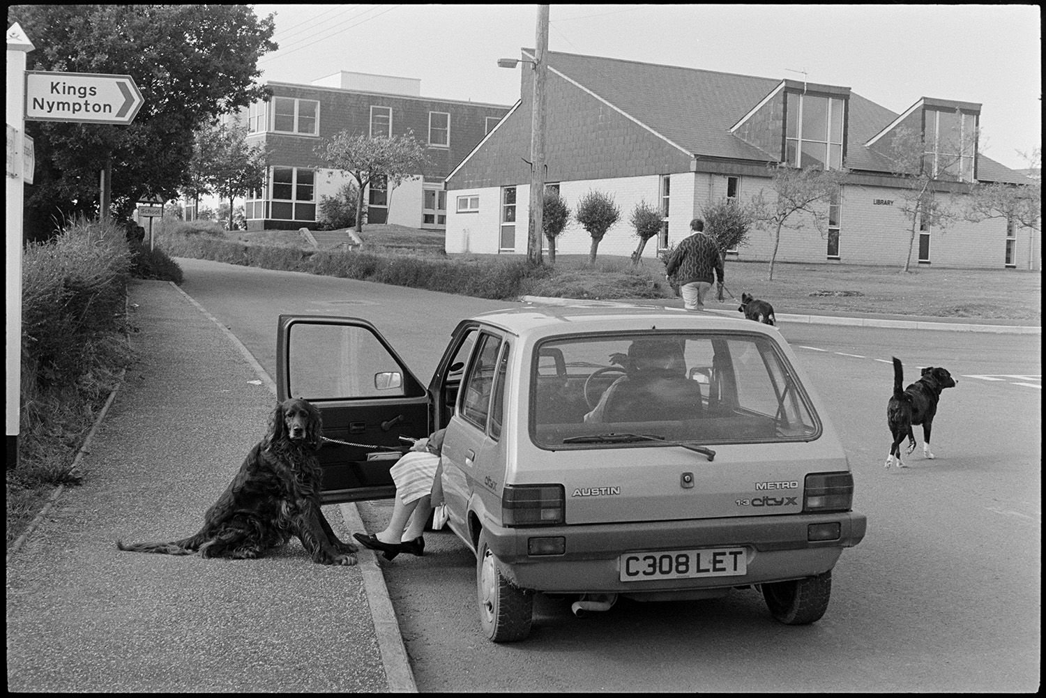 Street scenes with cars.
[A street scene showing South Molton Street, Chulmleigh. There is a Austin Metro car parked on the road with a woman passenger holding a red setter dog on a lead. A man is walking up the road with a dog on a lead, and another dog is crossing the road. A road sign pointing to Kings Nympton, and school buildings are visible in the background.]
