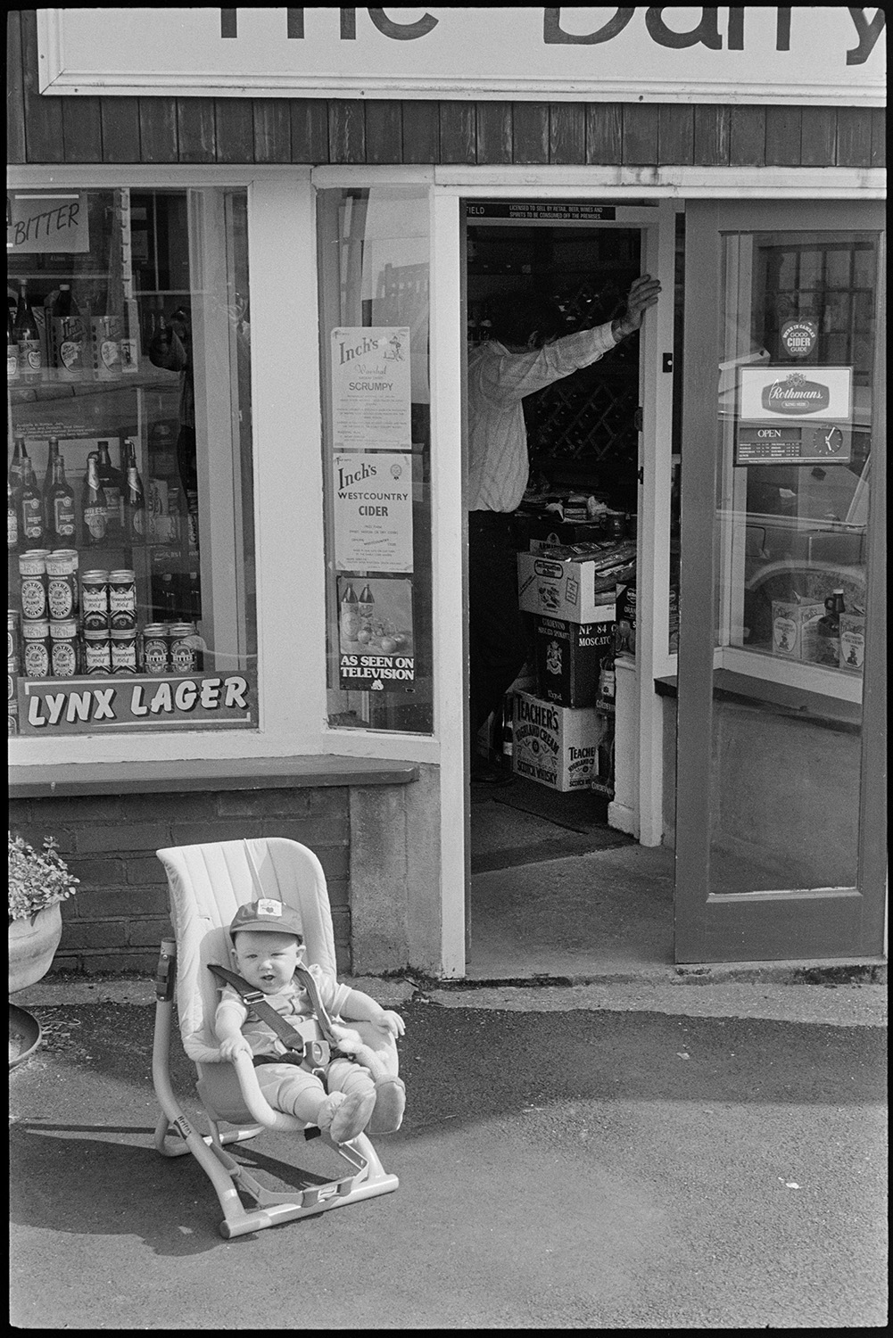 Baby in chair sitting outside shop.
[A young child sitting in a car seat outside The Dairy shop in Chulmleigh. Inch's Cider posters are displayed, with a window display of lager cans and bottles.]