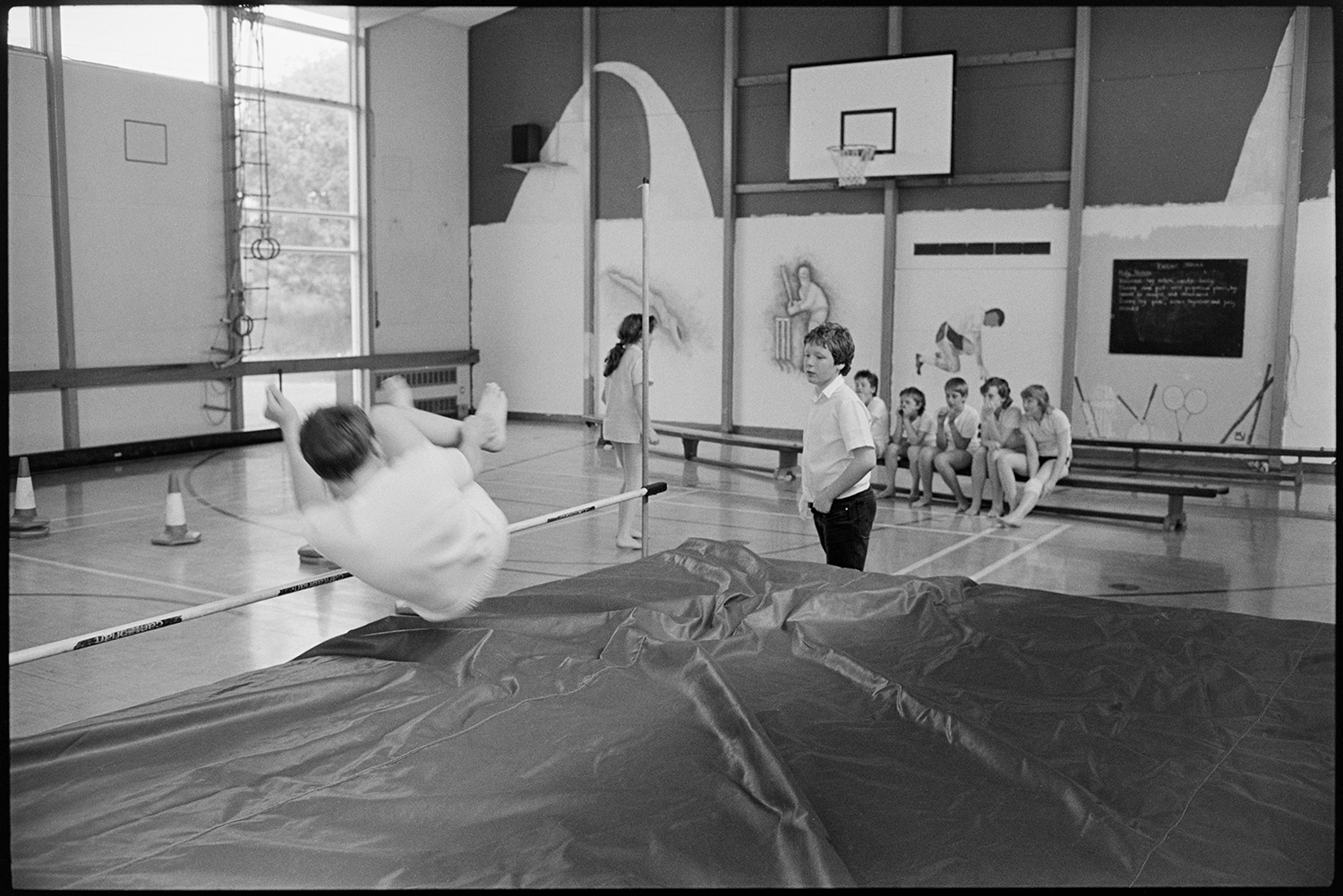 School lessons in classroom gymnastics, technology, craft, woodwork workshop.
[A gymnastics class at Chulmleigh Community College sports hall. A boy is completing the high jump, with another boy watching. There are pupils sitting on a bench in the background, and a rope ladder hanging from the ceiling.]