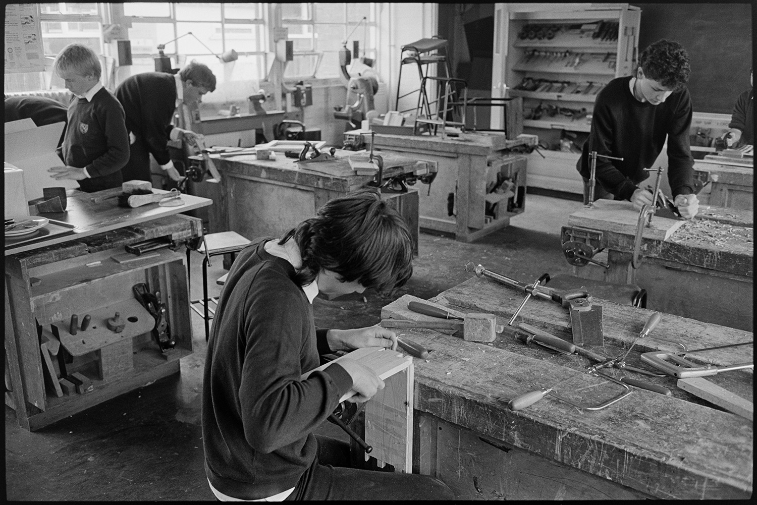 School lessons in classroom gymnastics, technology, craft, woodwork workshop.
[Woodworking class at Chulmleigh Community College. Four pupils are at benches working with tools including planes, clamps, saws, mallets and chisels. One pupil is planning a piece of wood which is clamped to the bench, and another is working on a piece with dovetail joints.]
