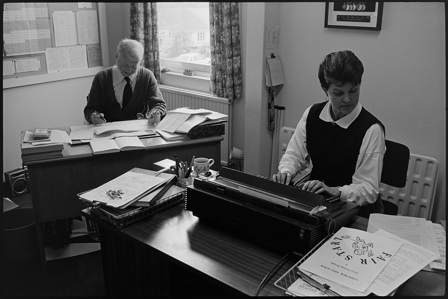 School lessons in classroom, examiners looking at art work. Secretary.
[Secretary working at her desk, using a typewriter, at Chulmleigh Community College. A man is also in the office working at another desk with various papers.]