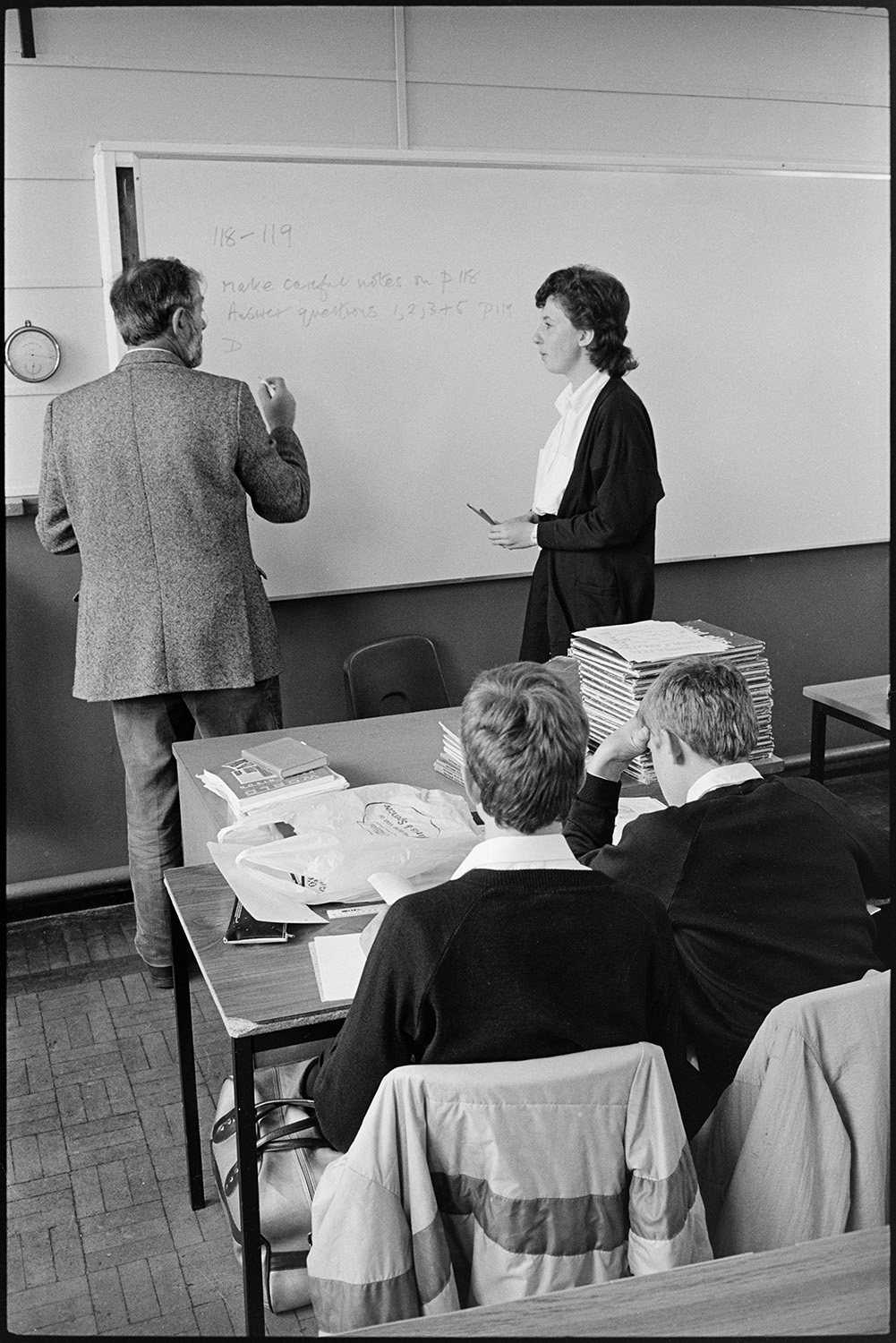 School lessons in classroom, examiners looking at art work. Secretary.
[A school teacher talking to a pupil while writing on a whiteboard at Chulmleigh Community College. There is a desk with a pile of books on it. Two pupils are sitting at their desk at the front of the class.]