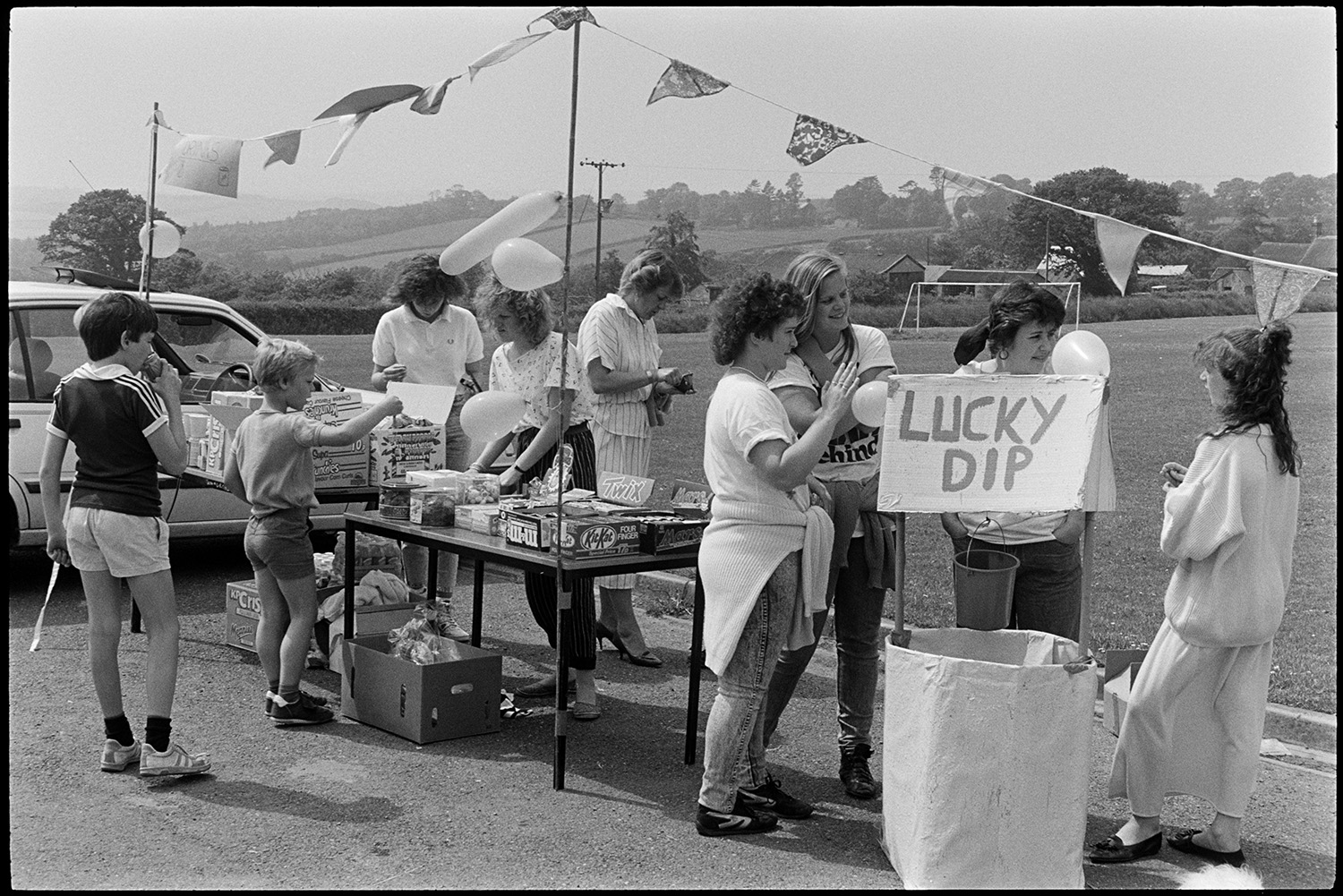 School Fete, stalls.
[A stall selling chocolates and snacks, alongside a Lucky Dip game, at the School Fete at Chulmleigh Community College. There are bunting and balloons around the stall, with the school playing fields and a wooded landscape in the background. Children and adults are gathered around and running the stalls.]
