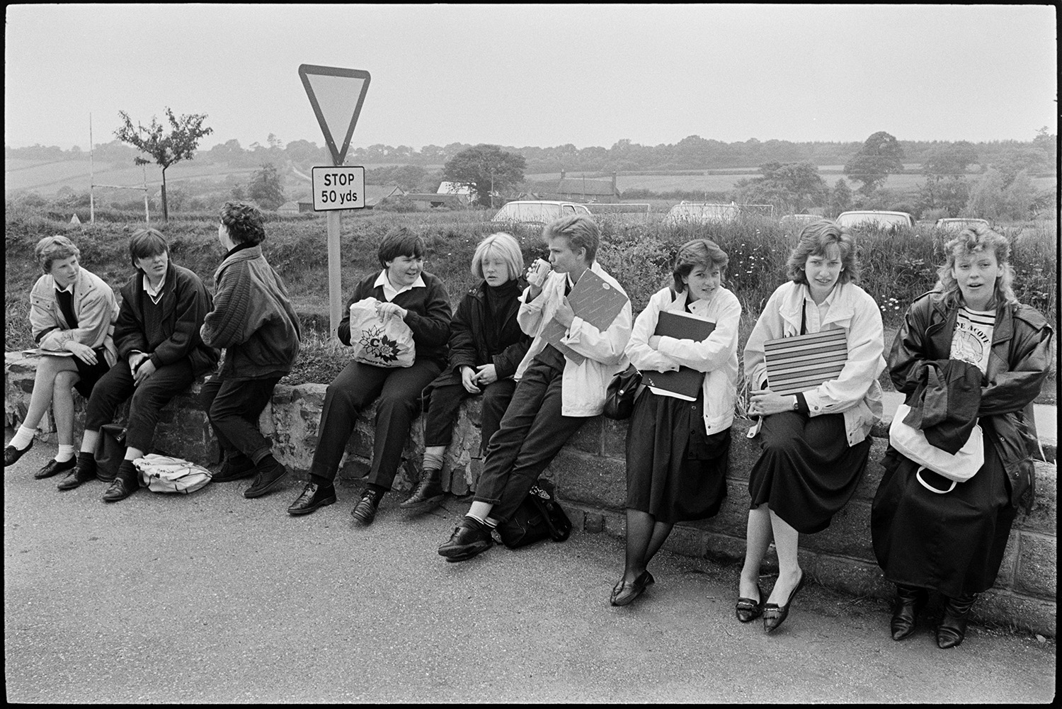 Schoolchildren waiting for bus after school and getting on to go home.
[Schoolchildren waiting for their bus after school at Chulmleigh Community College. They are sitting on a wall alongside a traffic warning sign, with a wooded landscape and parked cars in the background.]