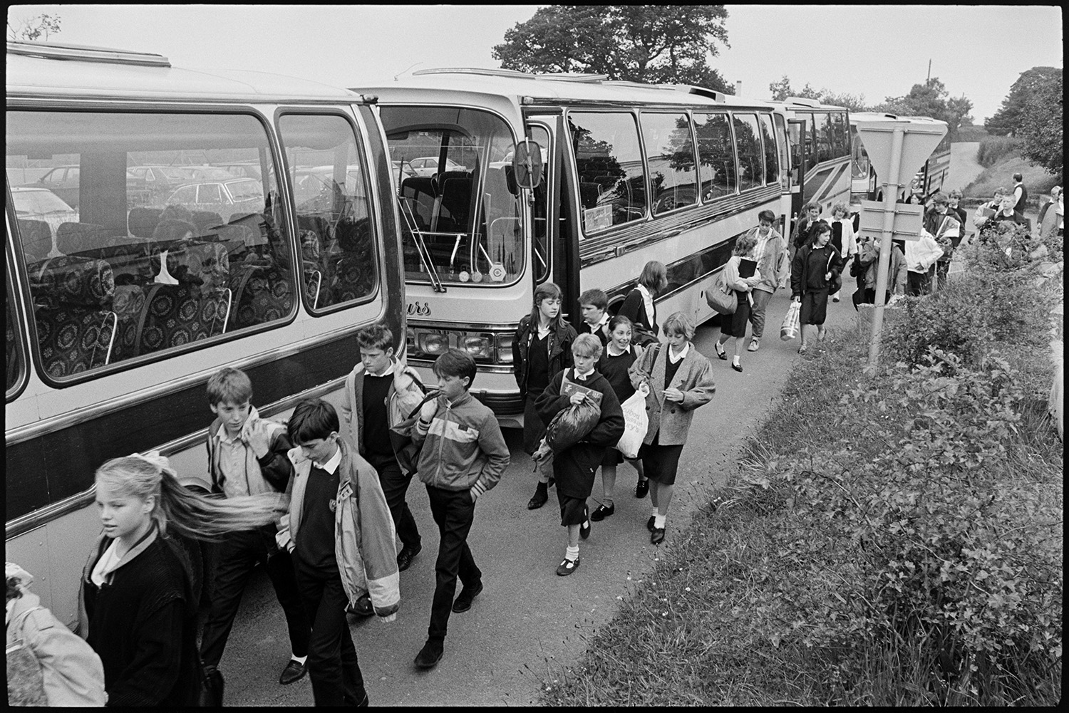 Schoolchildren waiting for bus after school and getting on to go home.
[Schoolchildren walking out to their buses to go home after school at Chulmleigh Community College. There is a row of five parked coaches on the road outside the school.]