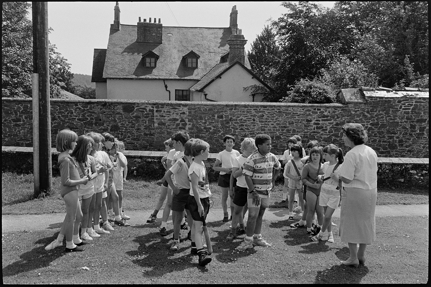 Children going through park to play rounders.
[Jean Harris, a teacher at Chulmleigh Primary School, with children lining up for games of rounders in Chulmleigh park. One child is holding a bat.]