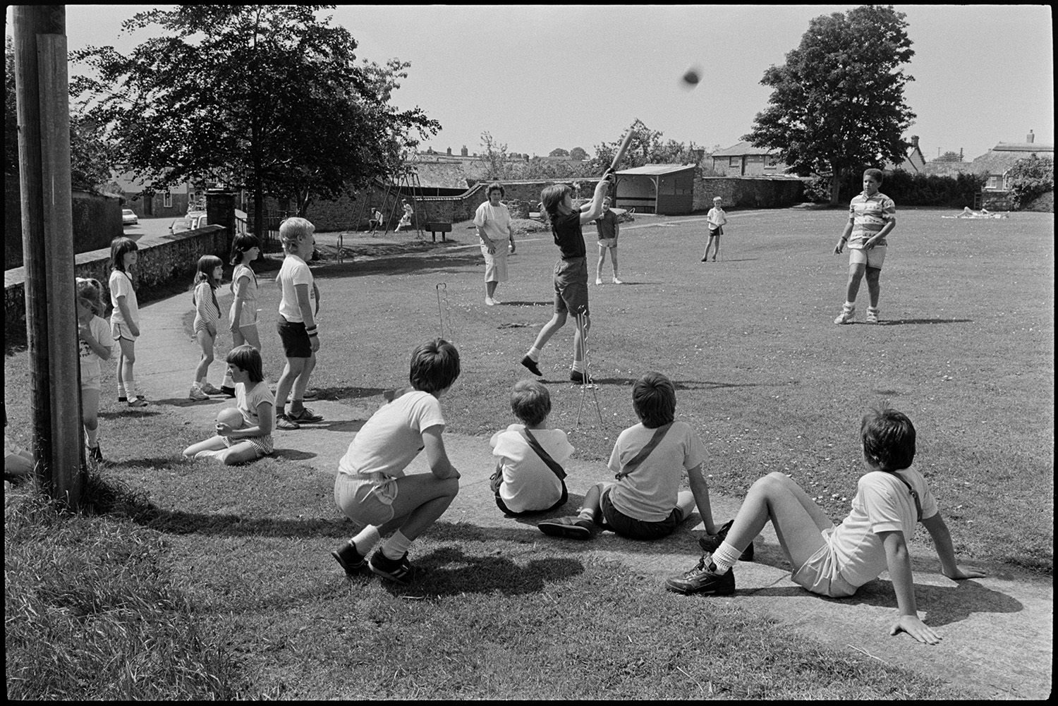 Children going through park to play rounders.
[Jean Harris supervising children from Chulmleigh Primary School playing rounders in Chulmleigh park. Some children are waiting to bat and some are sitting on the ground watching.]