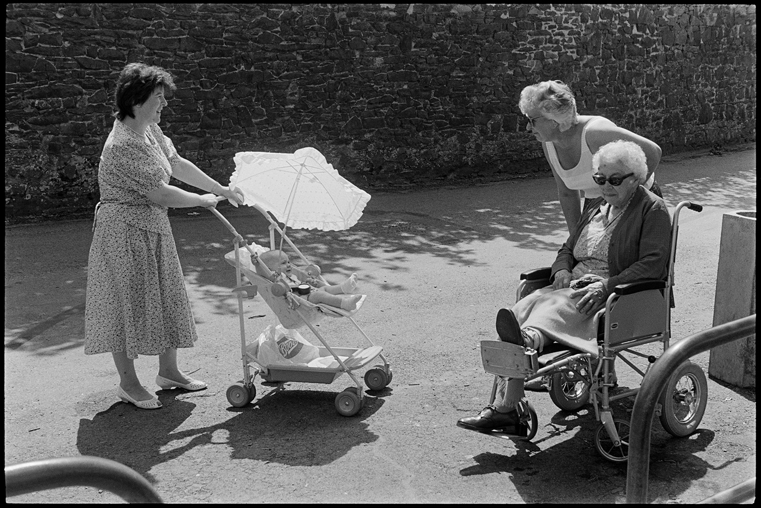 Woman mother and baby chatting to elderly woman in wheelchair.
[Chulmleigh street scene with two women, one in a wheelchair, talking to a woman with a baby in a pushchair.