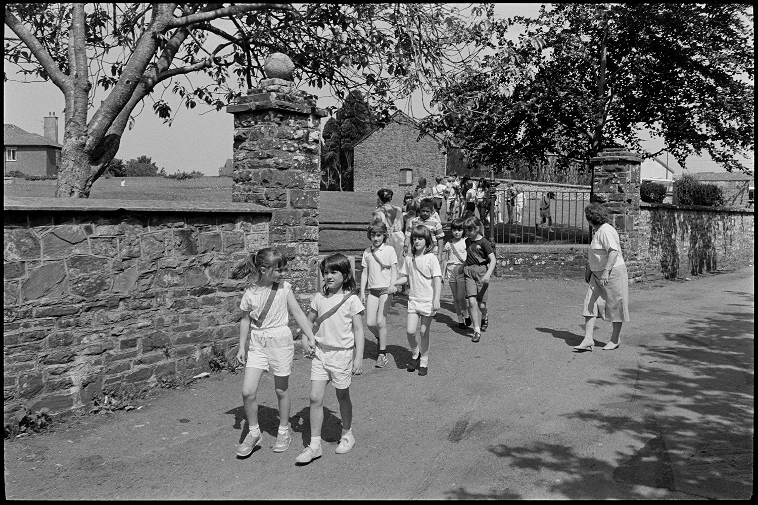 Children playing rounders in park. Returning through village.
[Children from Chulmleigh Primary School leaving Chulmleigh park and walking along a road back to school after playing rounders in the park. Jean Harris, a teacher, is supervising them.]