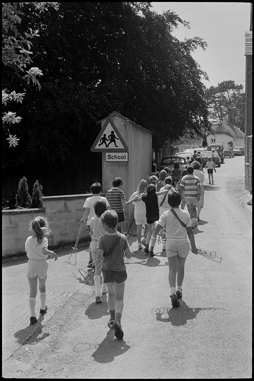 Children playing rounders in park. Returning through village.
[Children from Chulmleigh Primary School walking along a road past parked cars back to school after playing rounders in Chulmleigh park.]