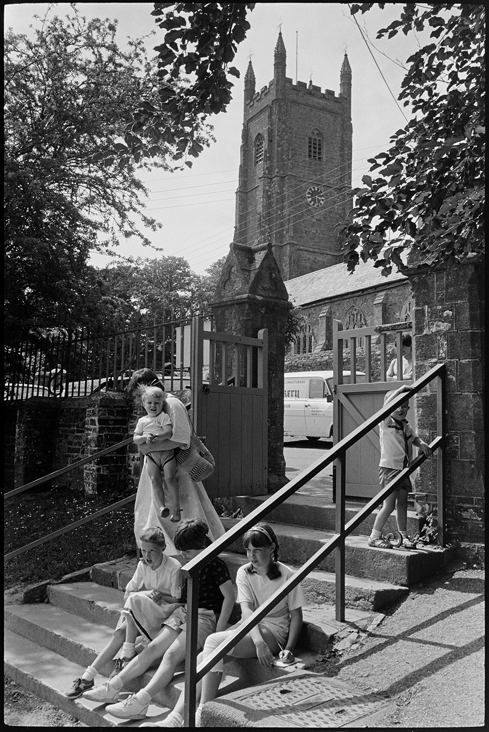 Children playing rounders in park. Returning through village.
[A mother carrying a young child, a boy playing by the hand rail and children sitting on the steps by the gate at Chulmleigh Primary School. Chulmleigh Church tower can be seen in the background.]