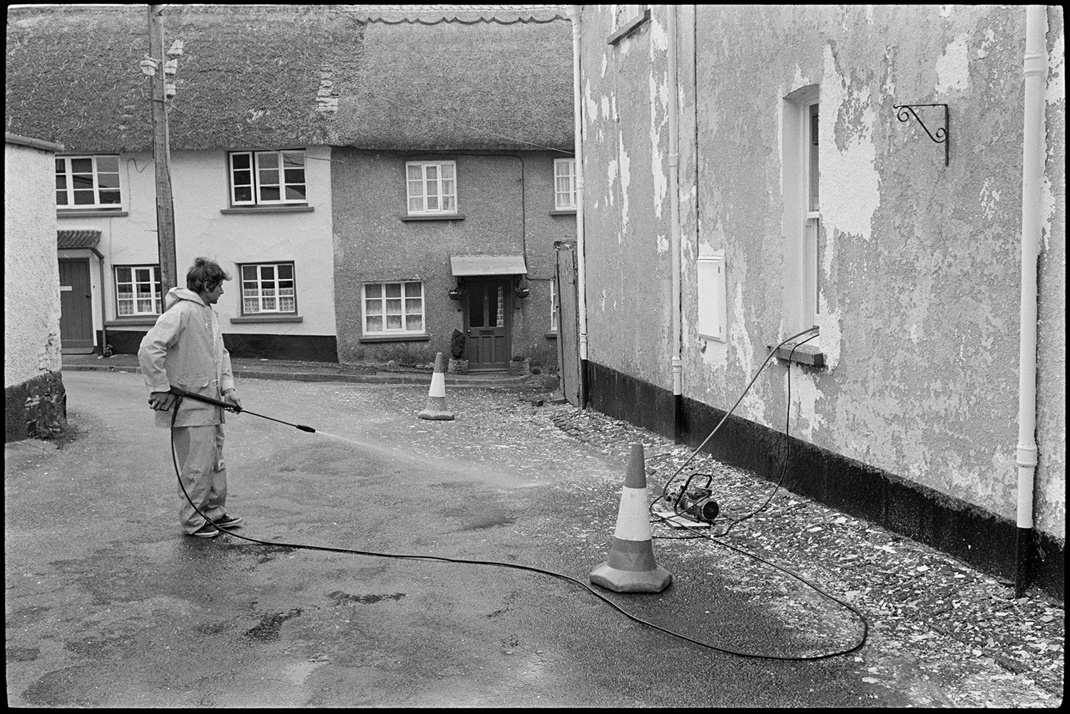 Man cleaning house with high pressure jet of water.
[A man dressed in waterproofs using a pressure washer to wash the walls of Peel House, Chulmleigh. The hose can be seen coming out of a ground floor window. Two thatched cottages are visible in the background.]