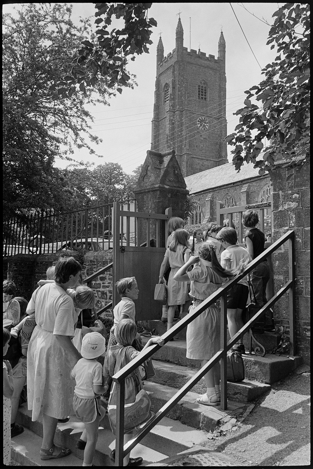Primary school. Children in playground and at school gates Church tower in background.
[A mother carrying a young child and a group of children walking up the steps to the gate at Chulmleigh Primary School. Chulmleigh church tower can be seen in the background.]