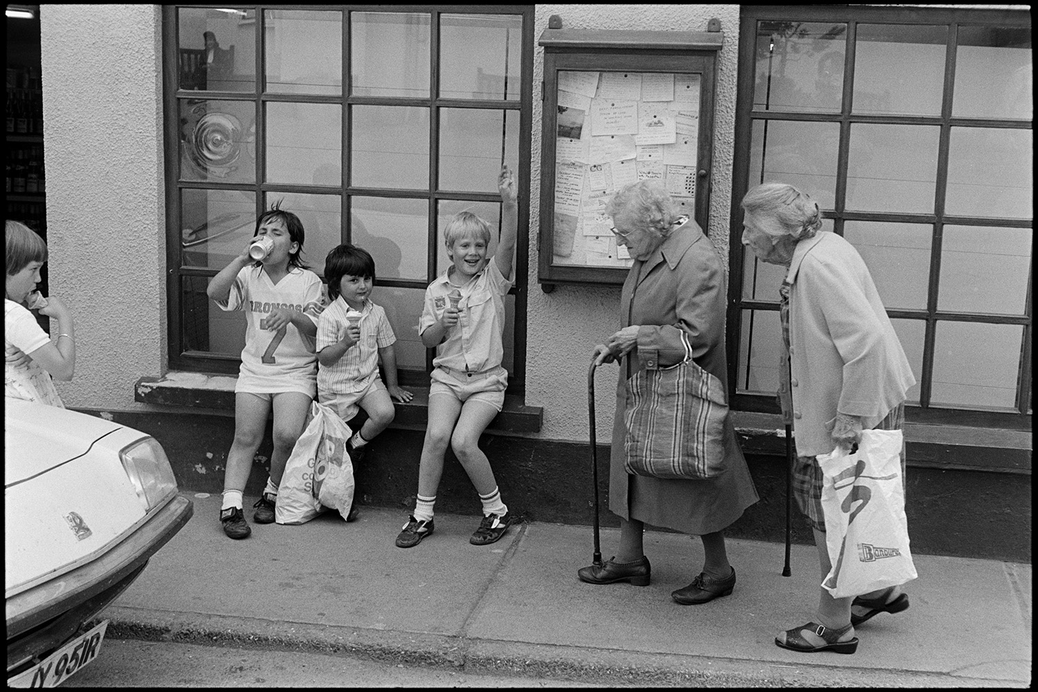 Street scenes with people and cars.
[Children sitting on a shop windowsill in a street in Chulmleigh with two women with walking sticks walking by. A notice board is on the wall in the background.]