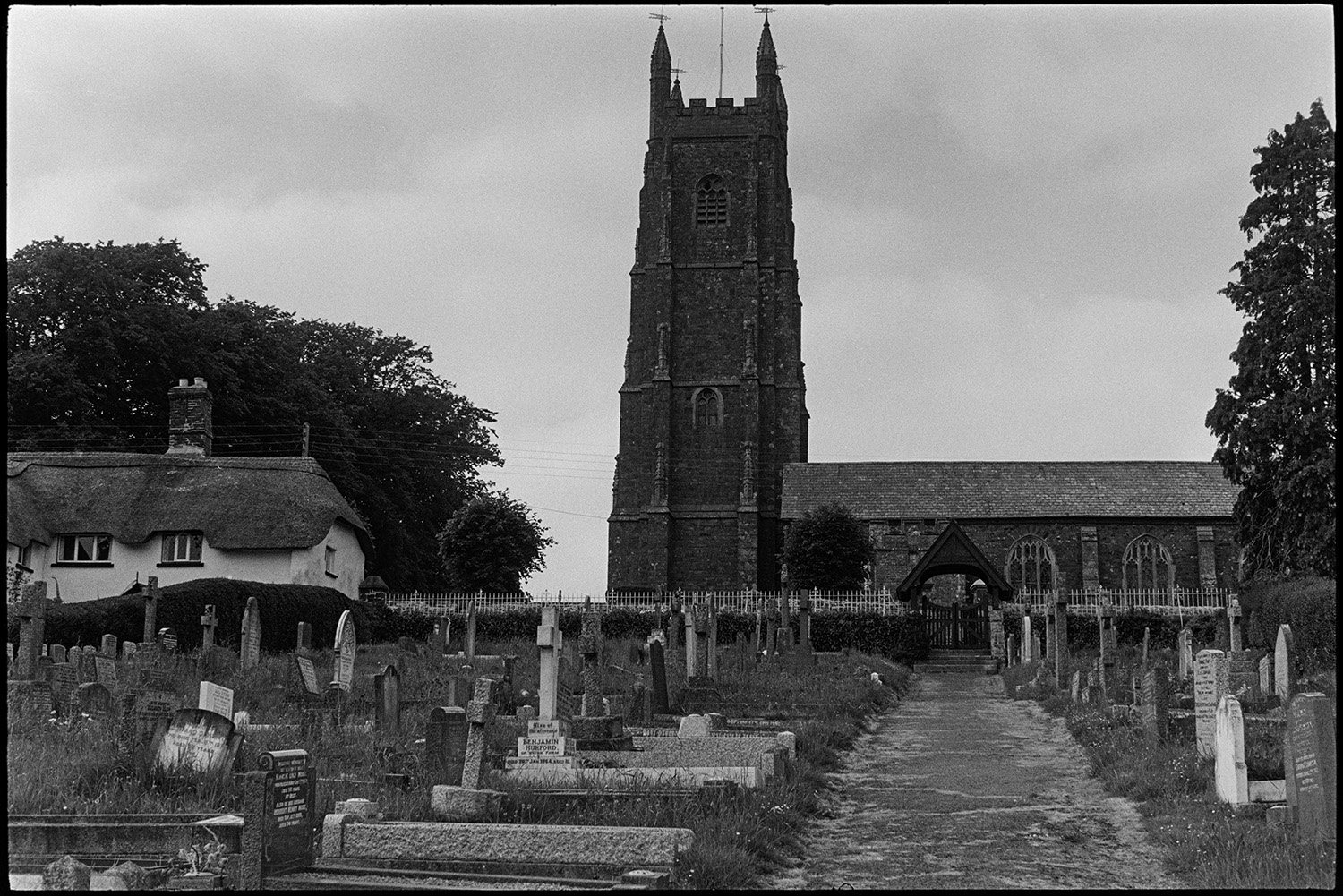 Street scenes with people and cars.
[A view of the church path with gravestones running up to Chulmleigh church and a thatched cottage, in Chulmleigh.]