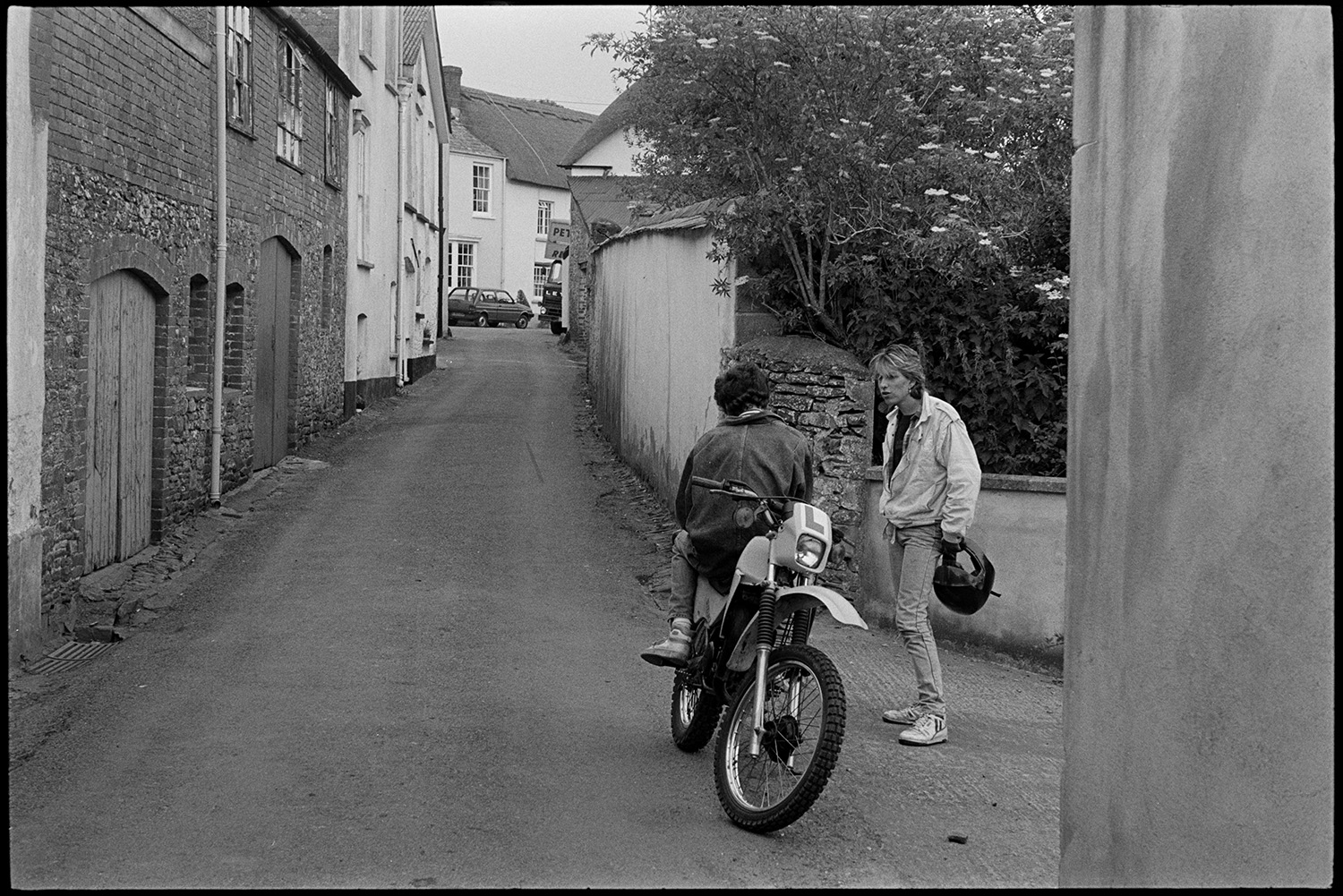 Street scenes with people and cars.
[Two young men talking in a narrow street in Chulmleigh. One is sitting backwards on a motorbike.]