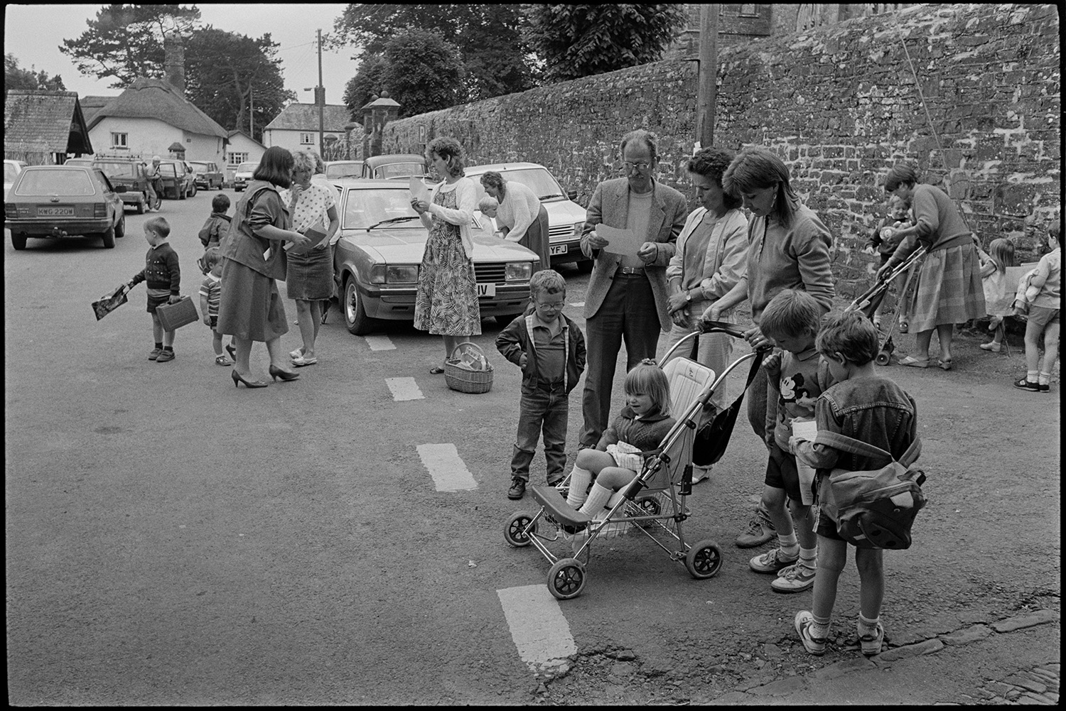 Women, mothers and children after school.
[Parents and children talking by a stone wall outside the school at Chulmleigh. Parked cars and cottages can be seen in the background.]
