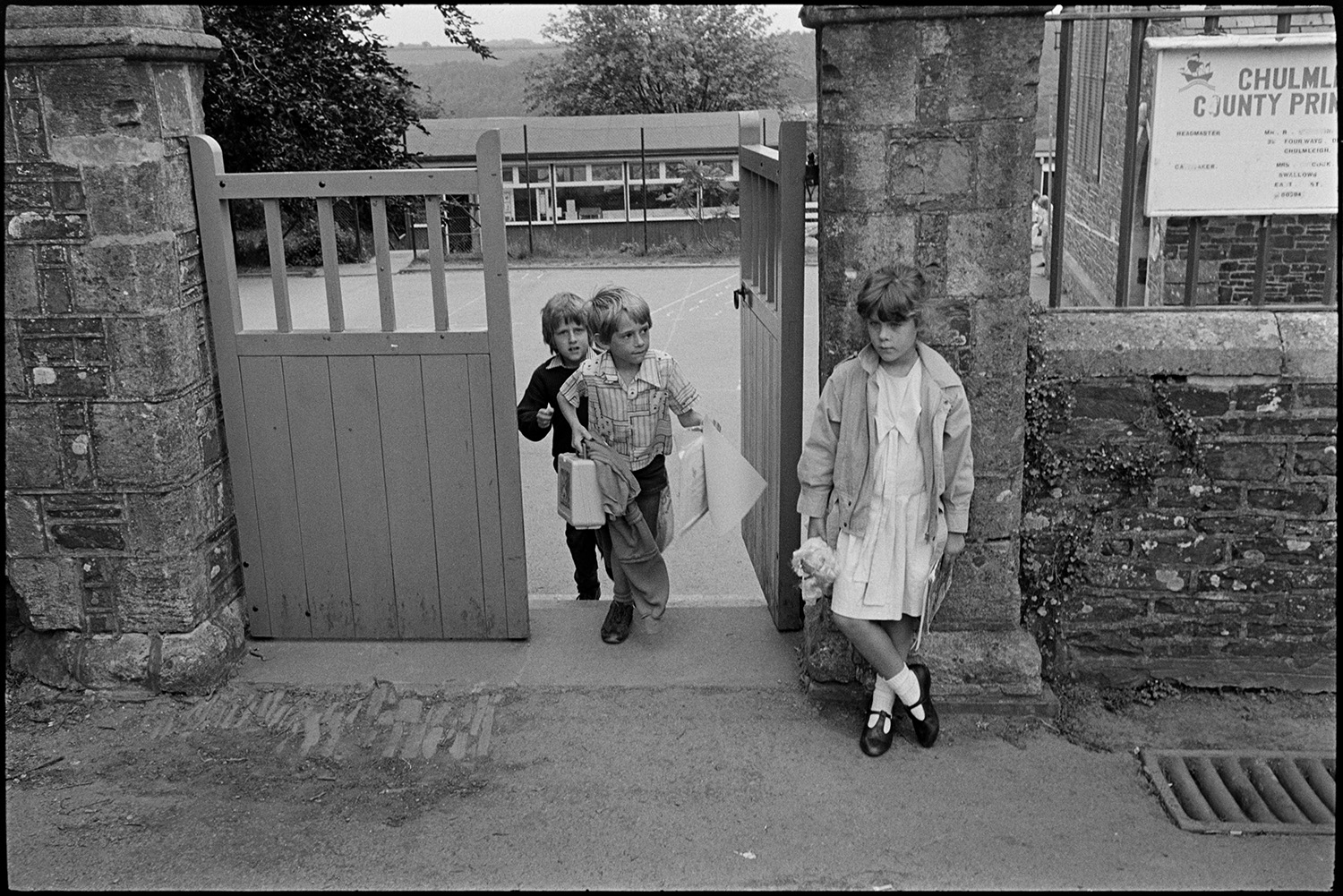 Women, mothers and children after school.
[Two boys leaving school and walking up the steps to the school gate at Chulmleigh Primary School. One of them is carrying pictures and a lunch box. A girl is standing by the wall outside the school.]