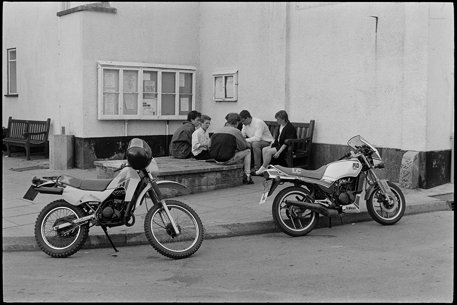 Street scenes with people and cars. 
[A group of young people sitting on a bench and wall in a street by The Red Lion Hotel in Chulmleigh. Two motorbikes are parked on the road in the foreground.]