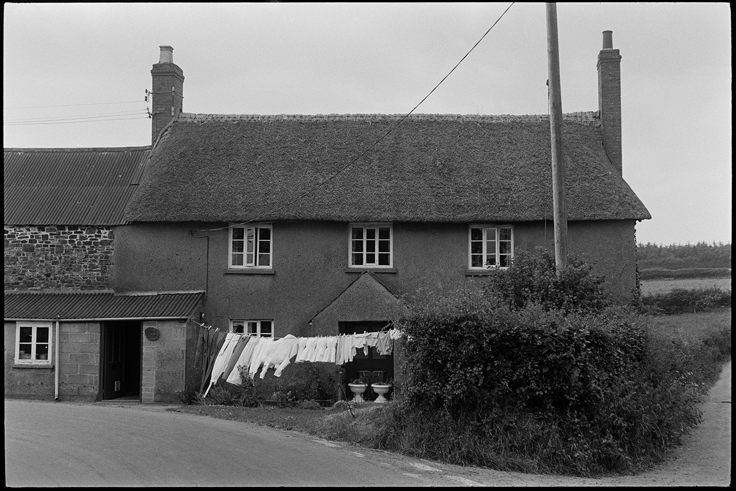 Cob and thatch farmhouse with clothes on washing line.
[Washing drying on a line at the front of Bonds, a cob and thatch farmhouse on a road junction at Chulmleigh. A stone barn with a corrugated iron roof is attached to the farmhouse.]