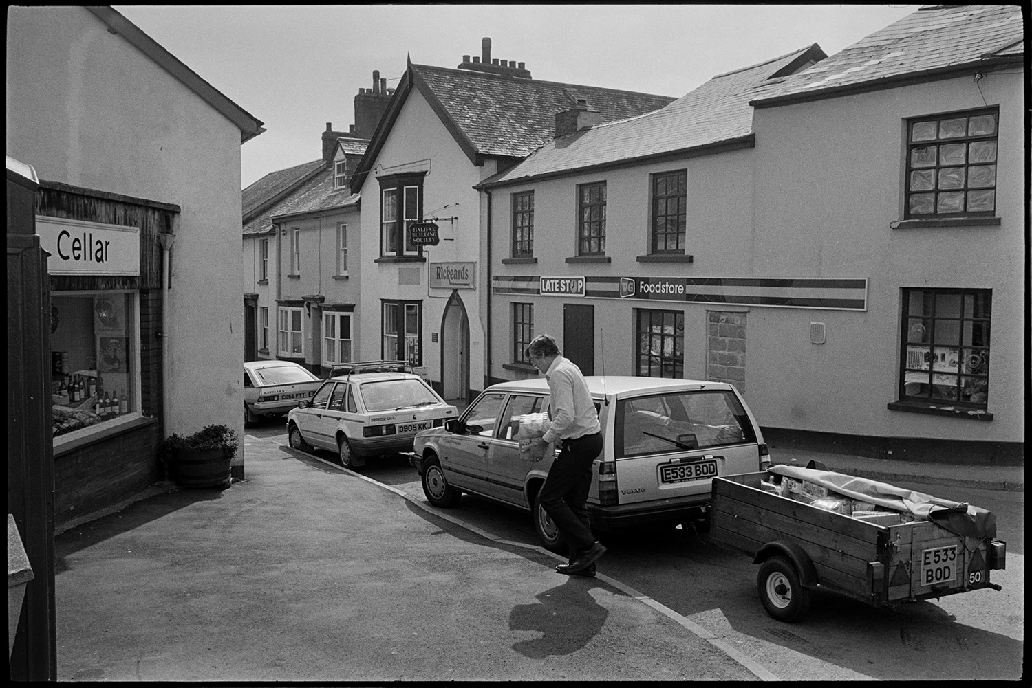 Street scenes with people and cars.
[A man unloading drinks from his parked car and trailer in a street in Chulmleigh. The shop fronts of Late Stop Food store, The dairy Cellar and Rickeards are visible in the background.]