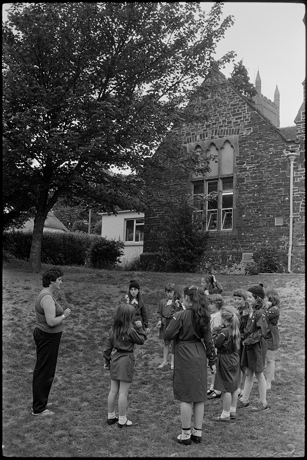 Brownies in school playground. Children in class setting off on nature walk on wood.
[A woman with a group of Brownies standing in a circle on grass in front of Chulmleigh Primary School.]