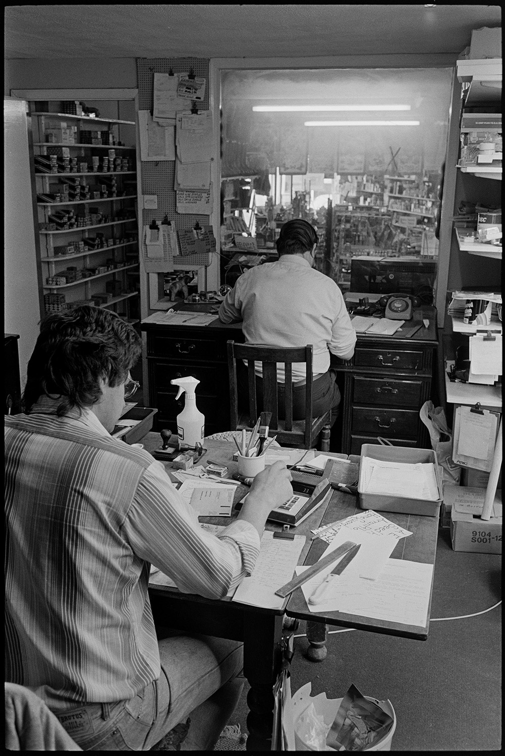 Interior of ironmongers shop. Man working at desk doing accounts. Fine dresser.
[Rear view of Tim Kingdon and Alec Brownscombe working in the back office of A E Kingdon ironmongers shop in Fore Street, Chulmleigh. The desks are covered with papers.]