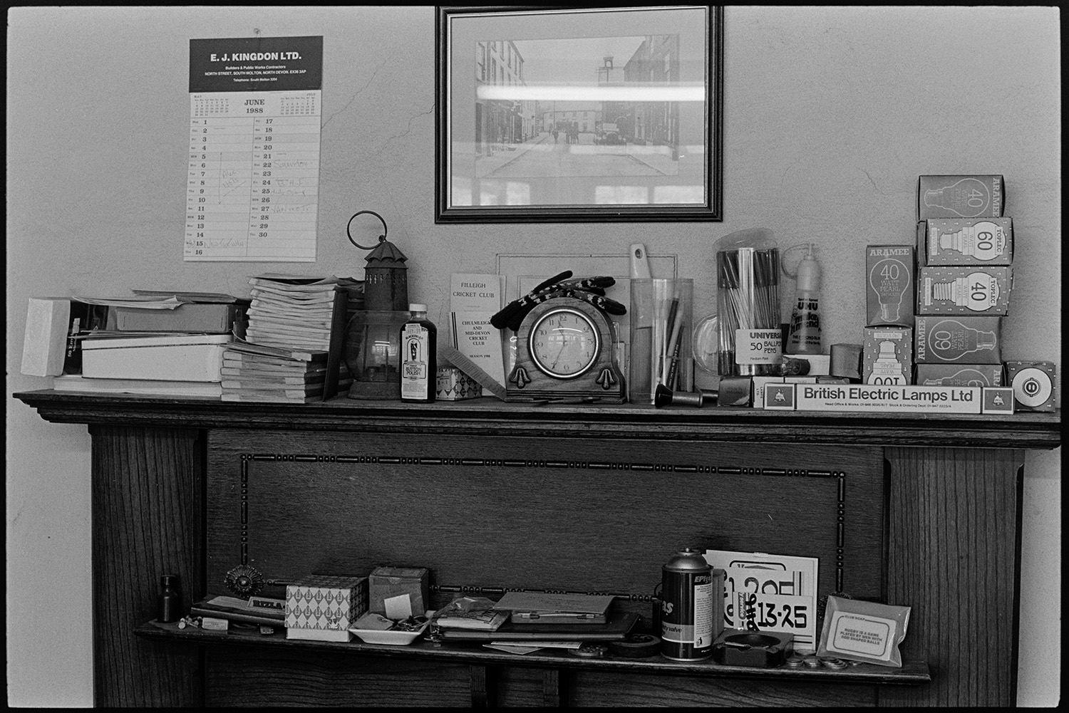 Interior of ironmongers shop. Man working at desk doing accounts. Fine dresser.
[Mantelpiece with clock, light bulbs, pens, a lamp and various other items at A E Kingdon ironmongers in Fore Street, Chulmleigh. A business calendar is hung on the wall.]