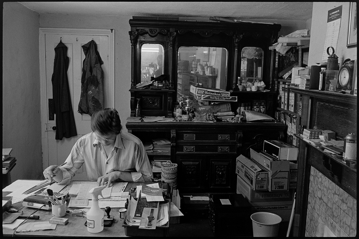 Interior of ironmongers shop. Man working at desk doing accounts. Fine dresser.
[Tim Kingdon working at his desk in the office at A E Kingdon ironmongers in Fore Street, Chulmleigh. A carved dresser and mantelpiece with various items, including files and boxes can be seen in the background.]