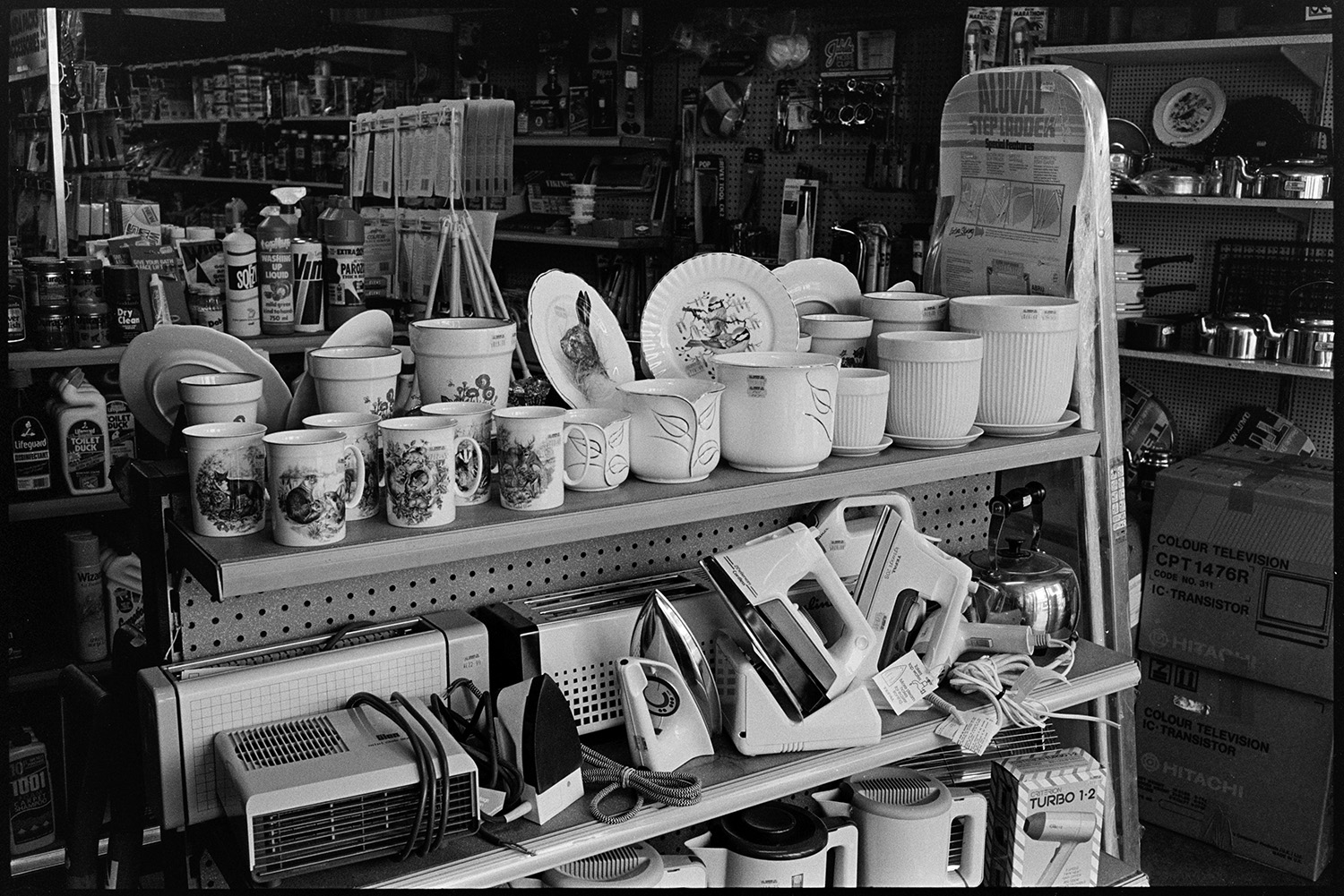 Interior of ironmongers shop. Assistant serving customer. Goods on shelves. Hardware.<br />
[Mugs, plant pots and electrical goods, including irons and kettles, displayed on shelves in A E Kingdon, ironmongers shop in Fore Street, Chulmleigh.]