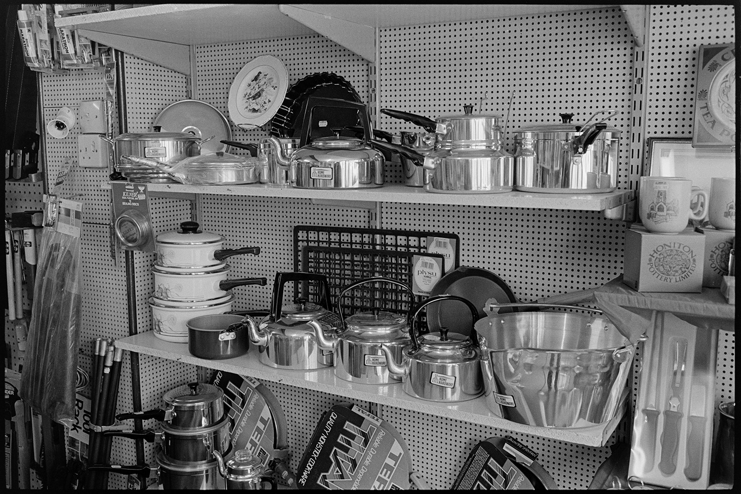Interior of ironmongers shop. Assistant serving customer. Goods on shelves. Hardware.<br />
[Saucepans and kettles for sale displayed on shelves in A E Kingdon, ironmongers shop in Fore Street, Chulmleigh.]