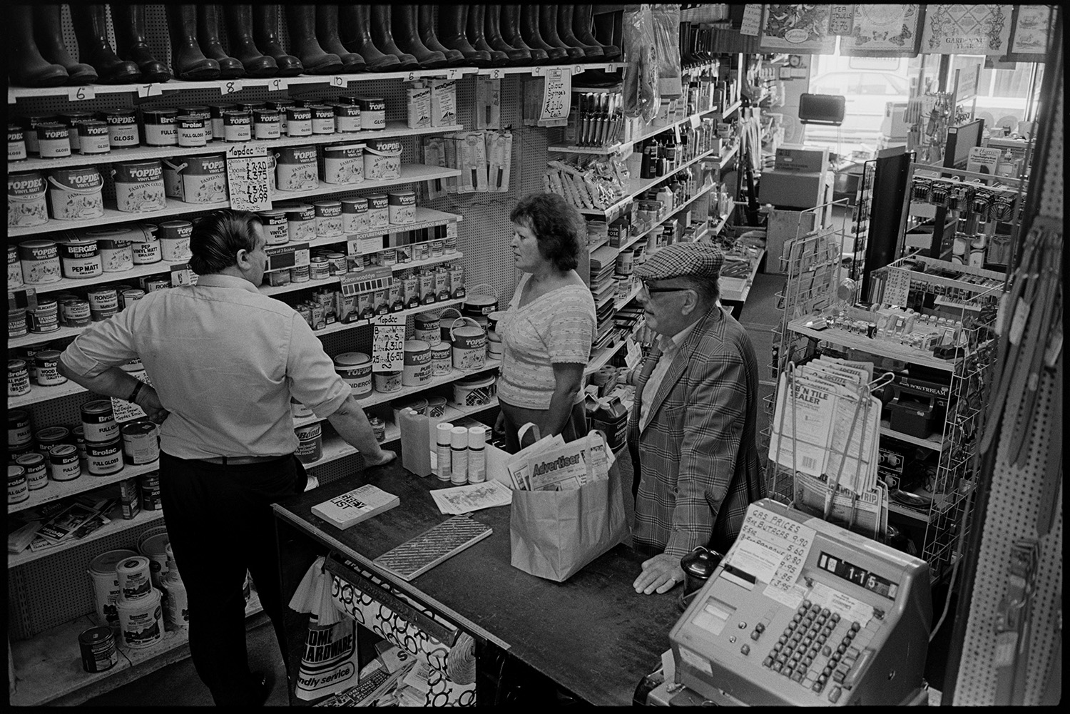 Interior of ironmongers shop. Assistant serving customer. Goods on shelves. Hardware.<br />
[A view of Alec Brownscombe serving customers at the counter of A E Kingdon ironmongers shop at Chulmleigh. Shelves are stocked with paint tins, Wellington boots and various other goods. The till can be seen on the shop counter.]