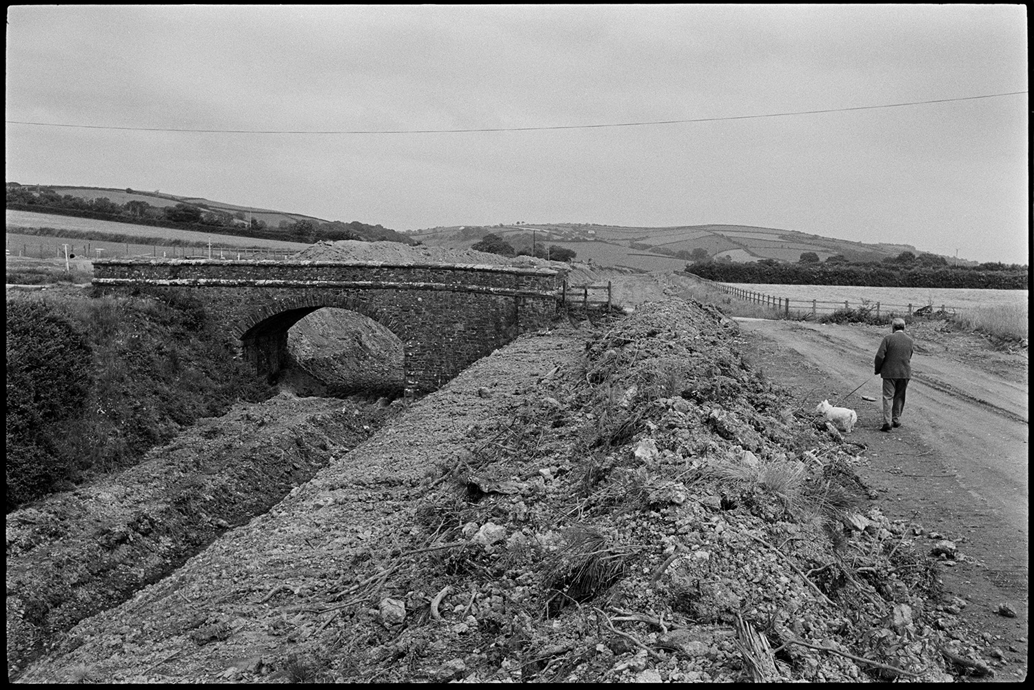 Site of new link road along old railway line. Stone bridge before demolition. Surveyor.<br />
[A man walking his dog next to the construction site, near South Molton, of the North Devon Link road, along an old railway line. The stone bridge was later demolished.]